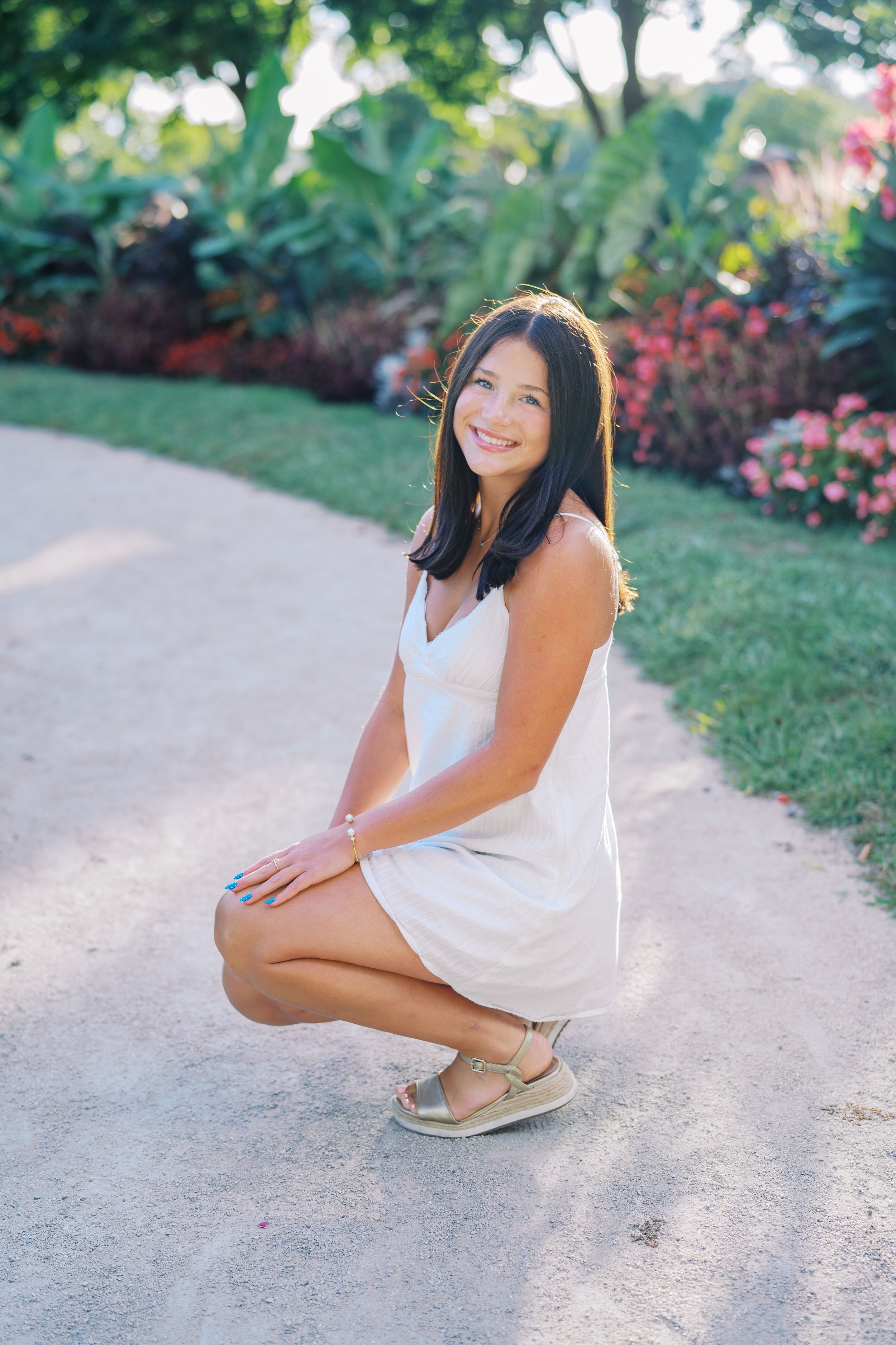 Person in white dress crouches on garden path with colorful flowers in background.