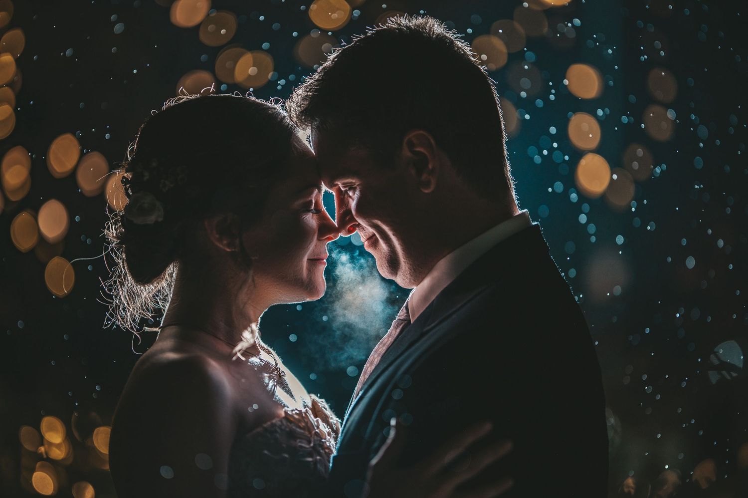 A romantic couple embraces in silhouette against a dark background with glowing bokeh lights creating a dreamy atmosphere.