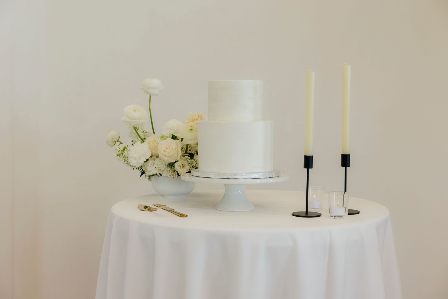 Elegant white two-tier wedding cake on stand with white floral arrangement and black taper candles on draped table.