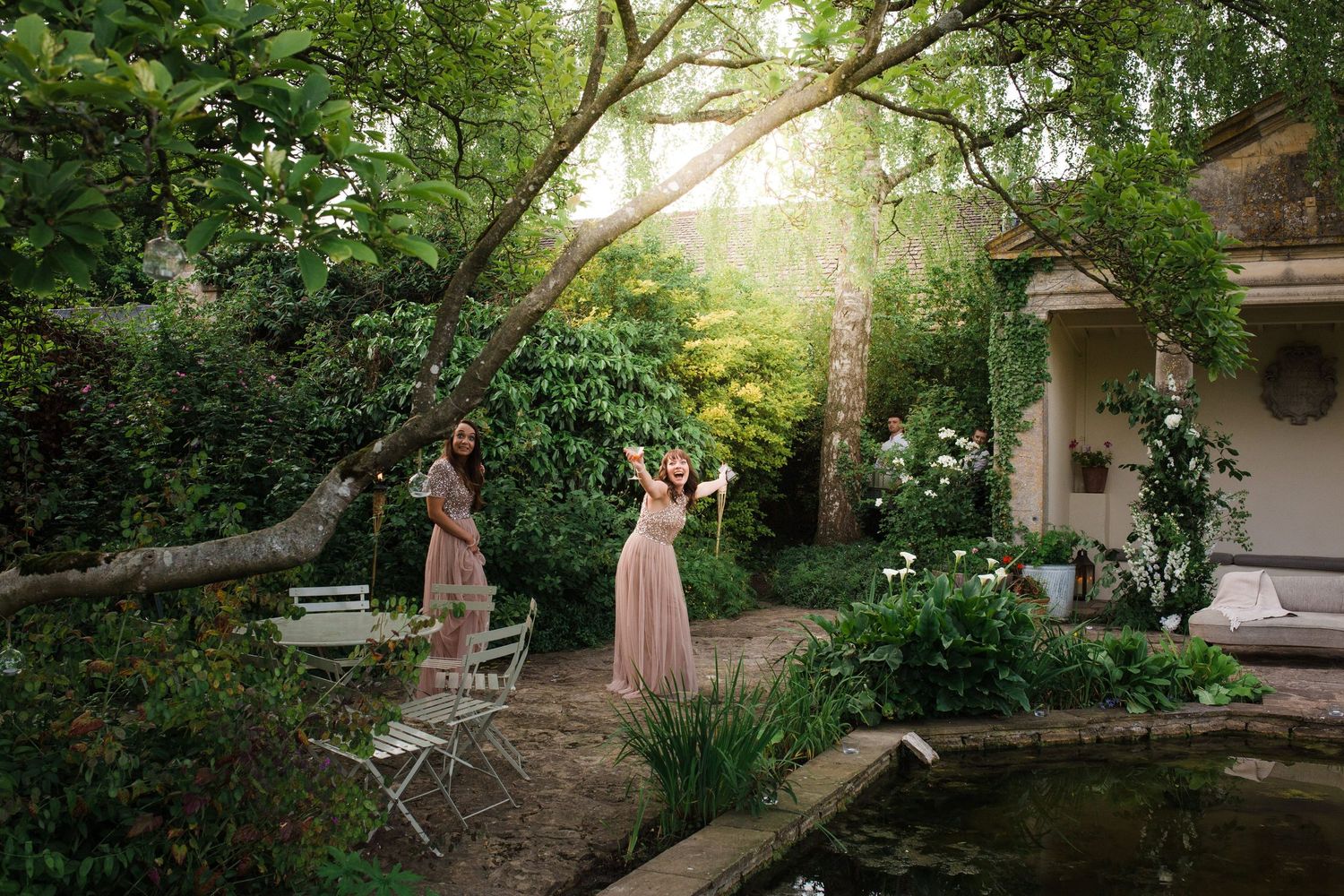 Two women in pink dresses stand in a lush garden near a pond, surrounded by greenery and soft natural light.