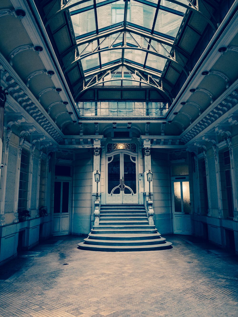 Grand abandoned hall with glass ceiling, ornate columns, and elegant staircase leading to arched doorway.