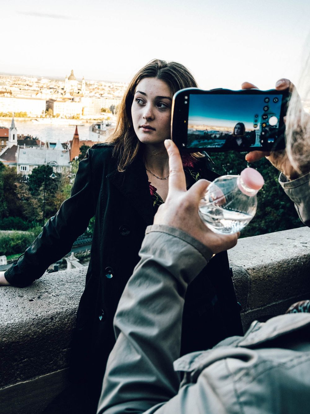 Young woman being photographed on a smartphone with a cityscape view in the background outdoors.