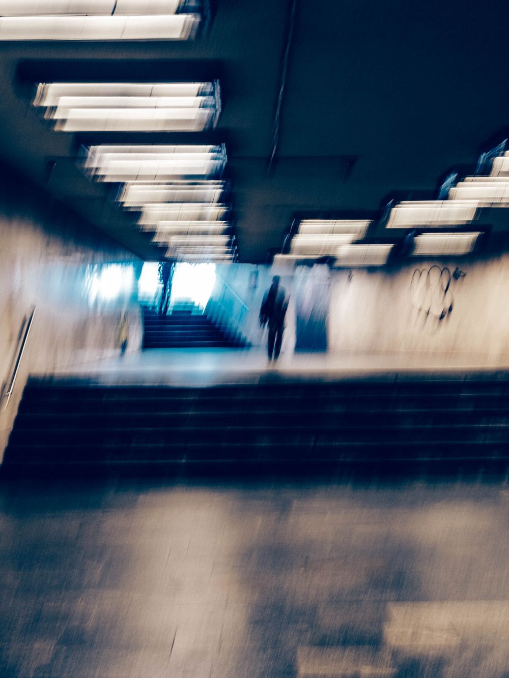 Blurry motion shot of a lone figure ascending subway stairs under fluorescent lights with graffiti on walls.