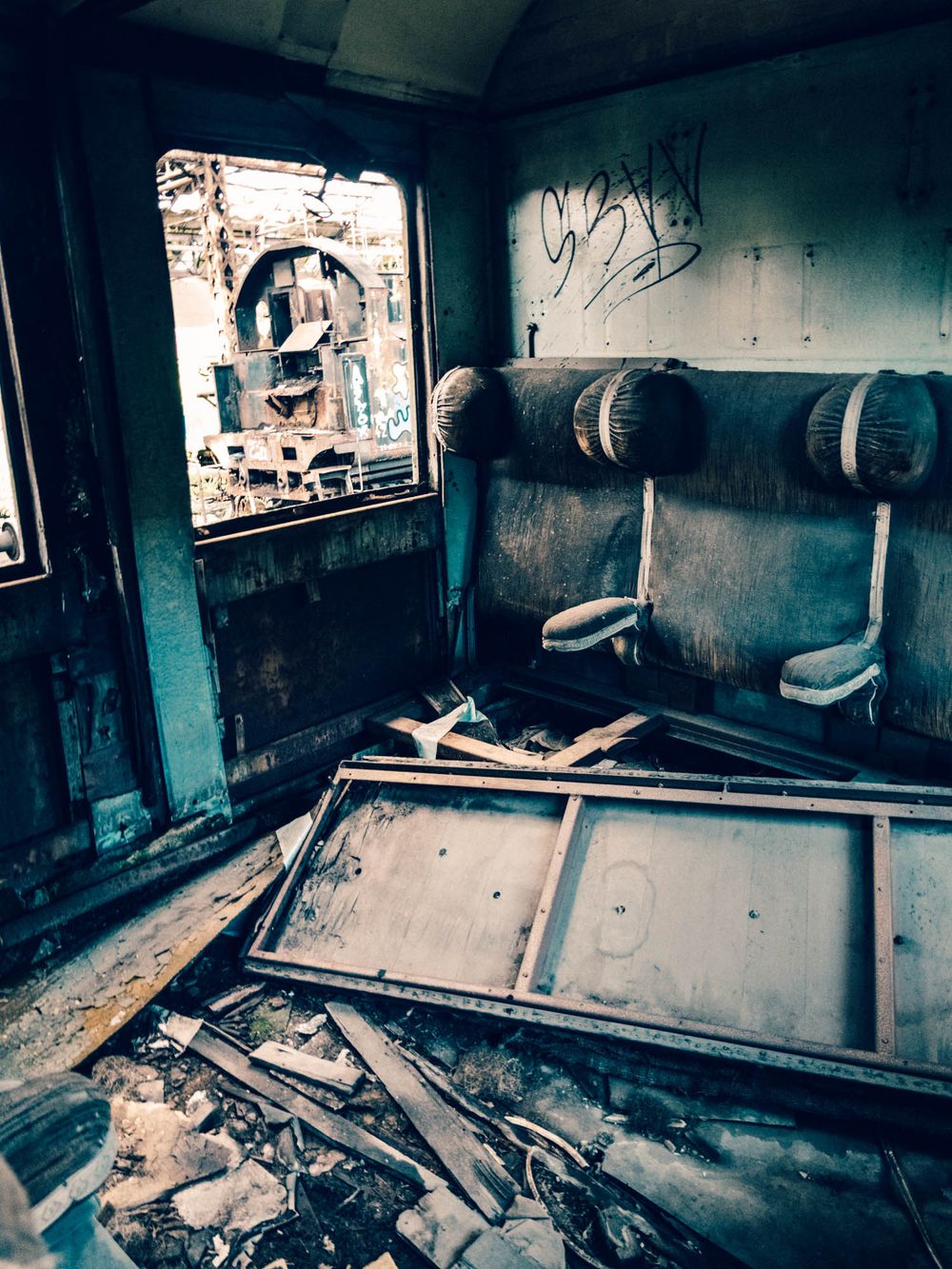 Abandoned train car interior with decayed seats, graffiti, debris, and broken wood panels in a derelict urban setting.