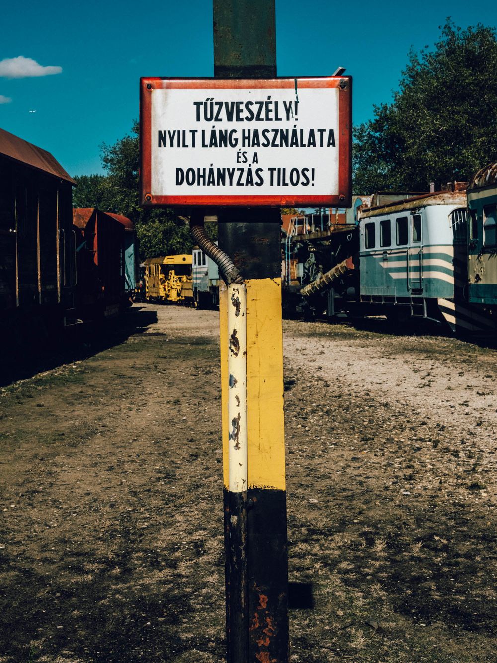 Hungarian fire hazard warning sign prohibiting open flames and smoking, mounted on yellow post at a railway yard.