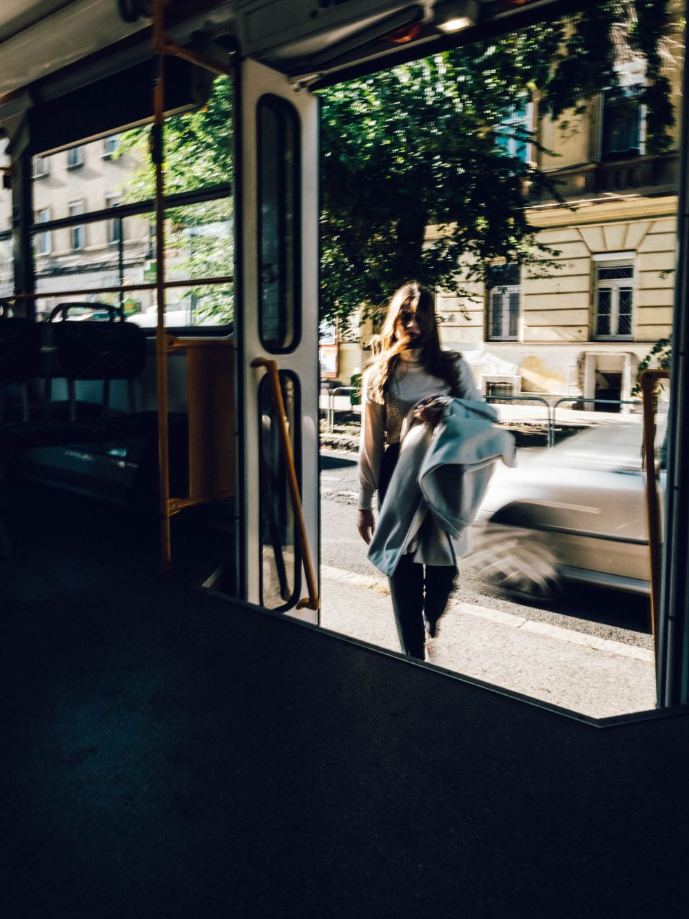 Woman boarding a city tram on a sunny urban street, carrying a gray blanket while checking her phone.