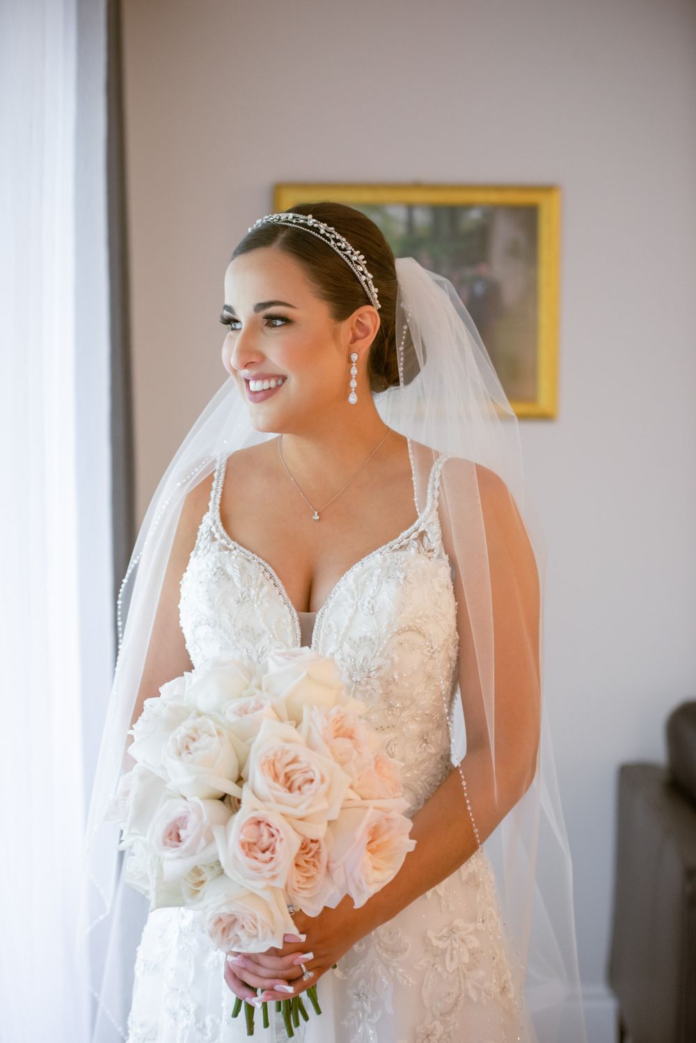 Radiant bride in lace wedding dress holds blush rose bouquet while smiling near a window.