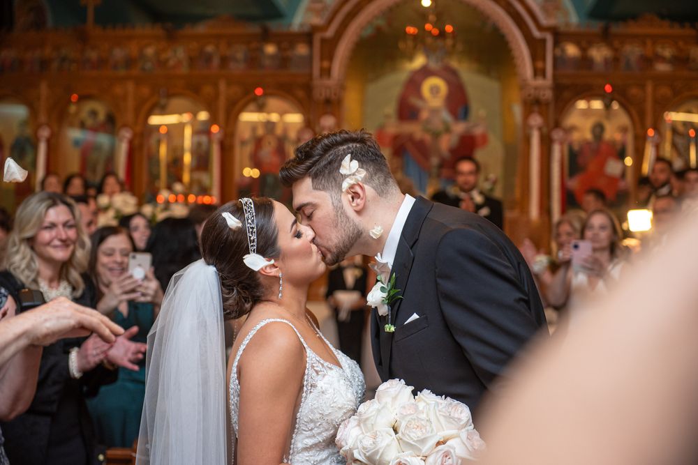 Bride and groom share a kiss in Orthodox church surrounded by guests after ceremony.