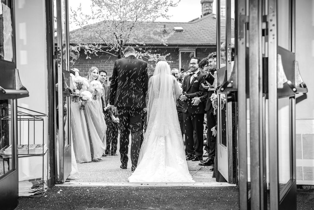 Black and white photo of bride and groom exiting church ceremony while guests celebrate outside.