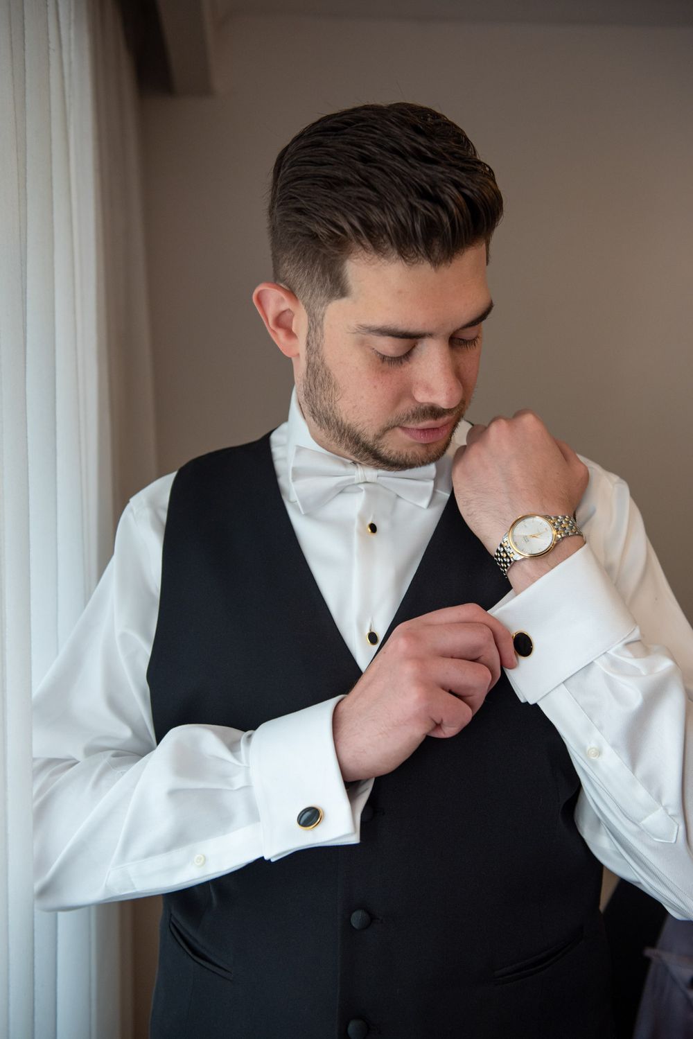 Dapper groom in black vest and white shirt fastening cufflinks before wedding ceremony.