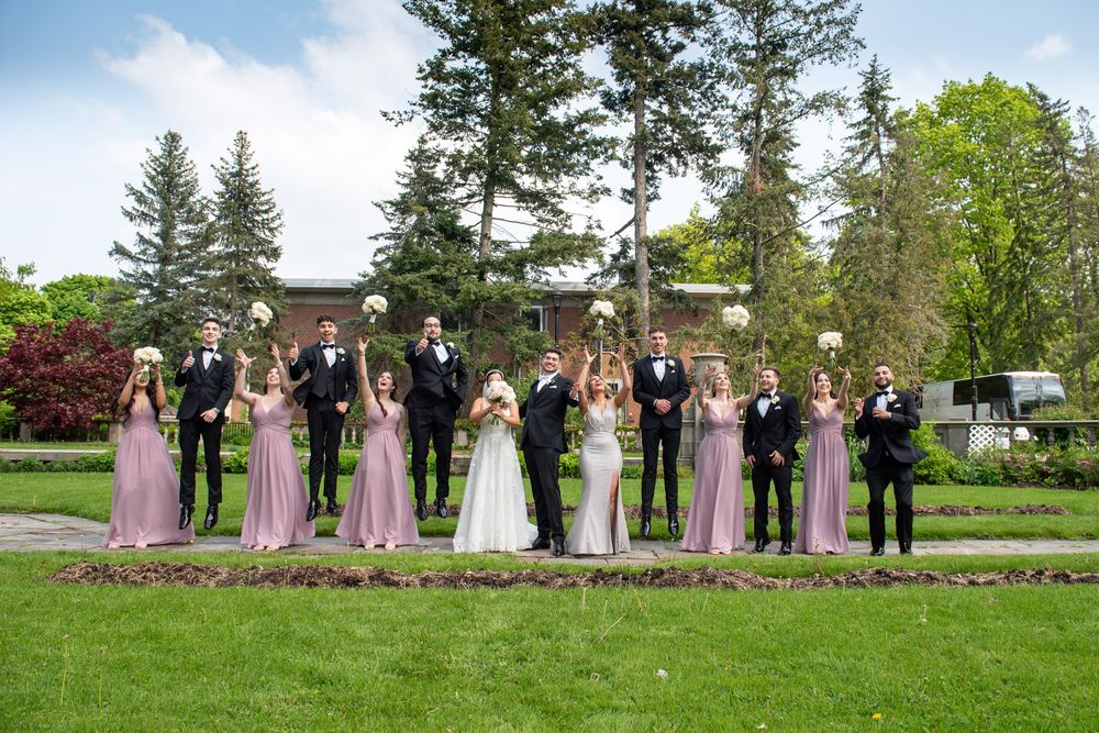 Wedding party in pink bridesmaid dresses and black tuxedos pose outdoors on green lawn with bouquets.