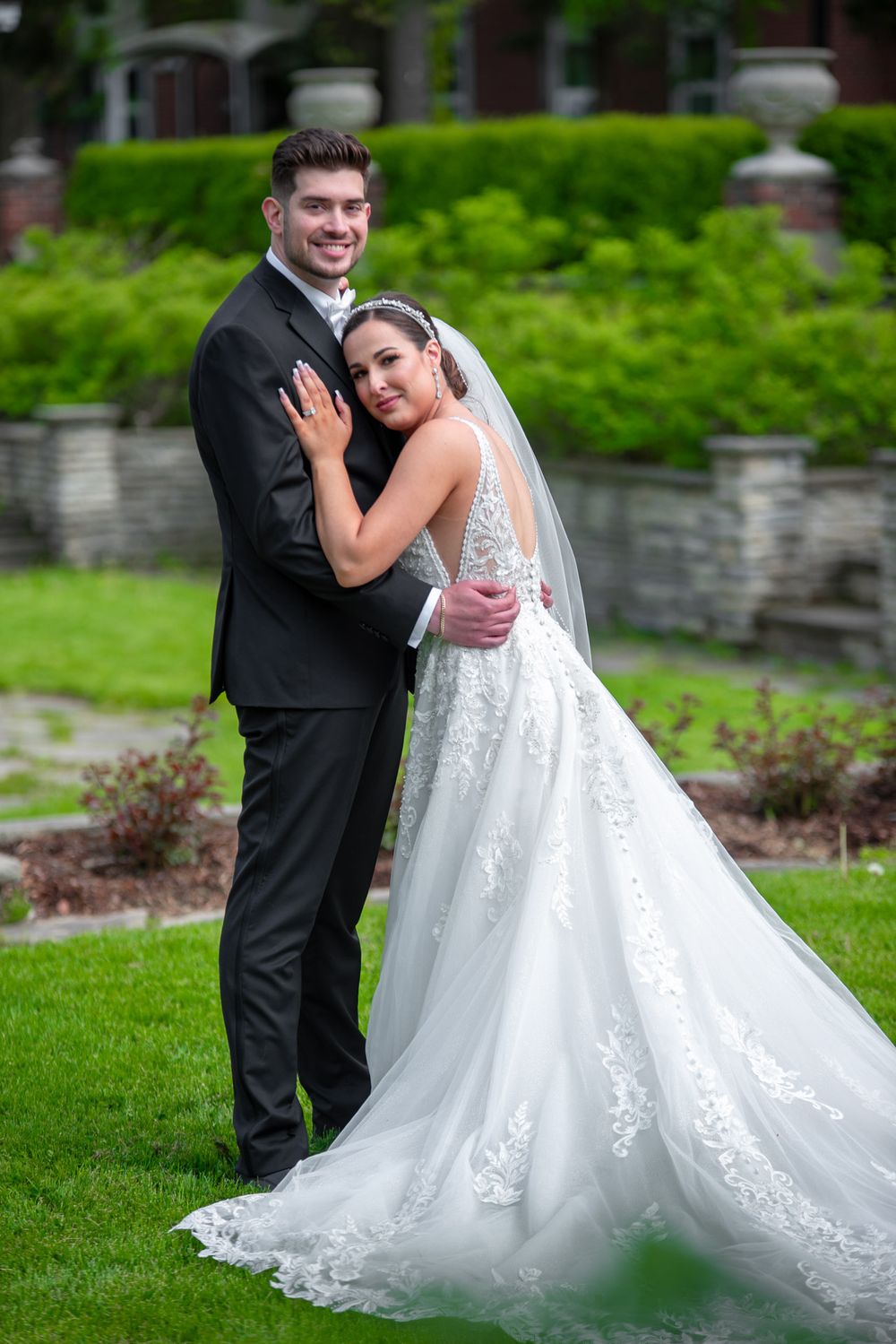 Smiling groom in black suit holds bride in lace wedding dress with train in a garden setting.