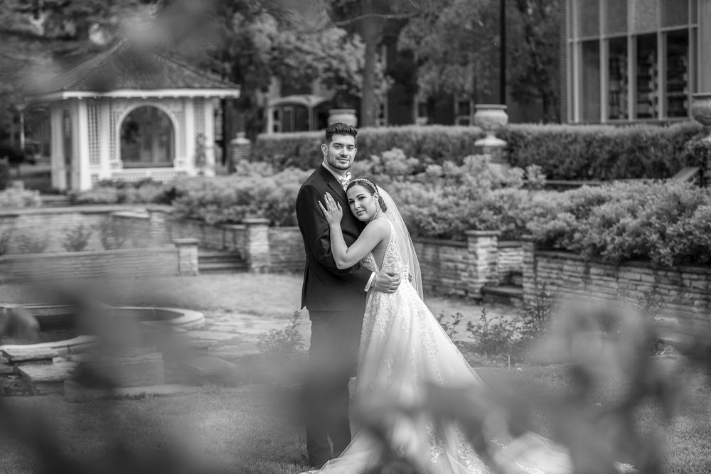Black and white photo of bride and groom embracing in an elegant garden with gazebo backdrop.
