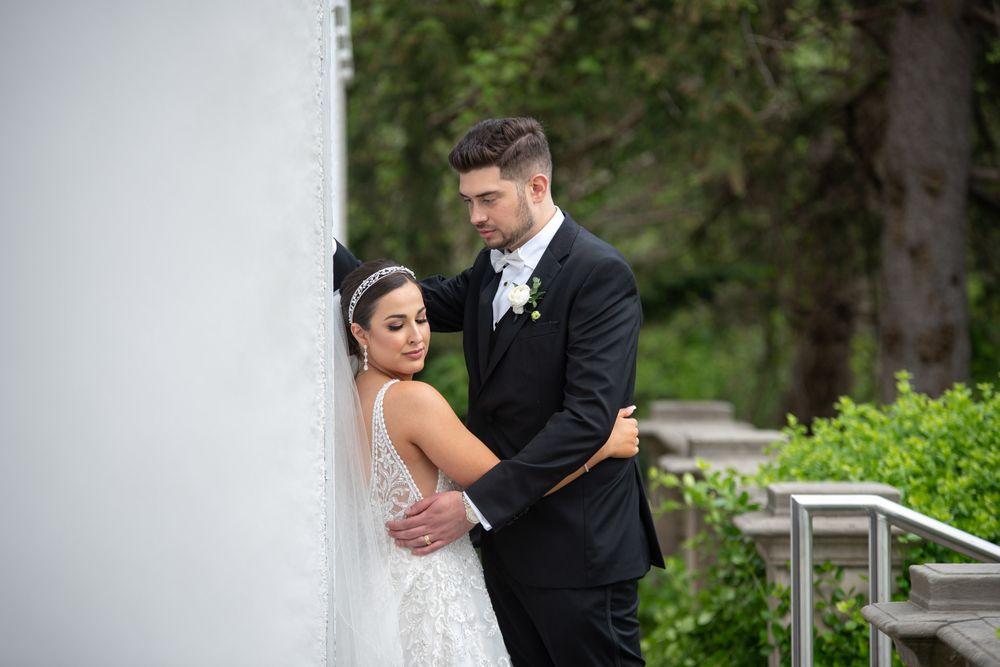 Elegant bride in lace gown and groom in black tuxedo posing together against a white wall outdoors.