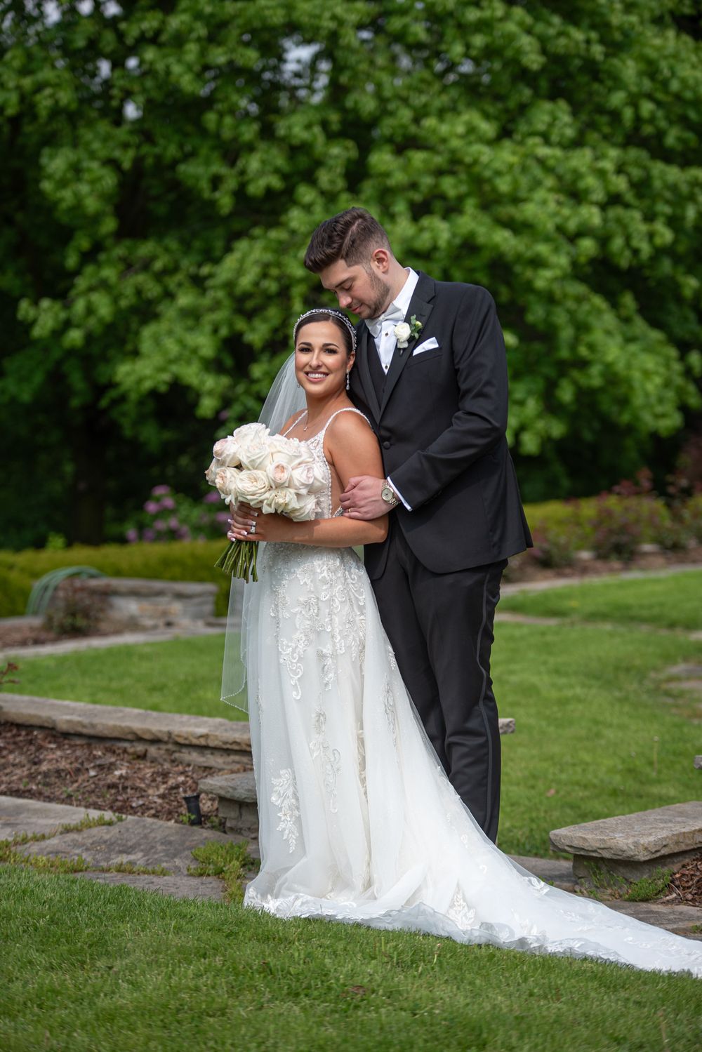 Bride smiles holding white rose bouquet as groom embraces her from behind in a green garden.