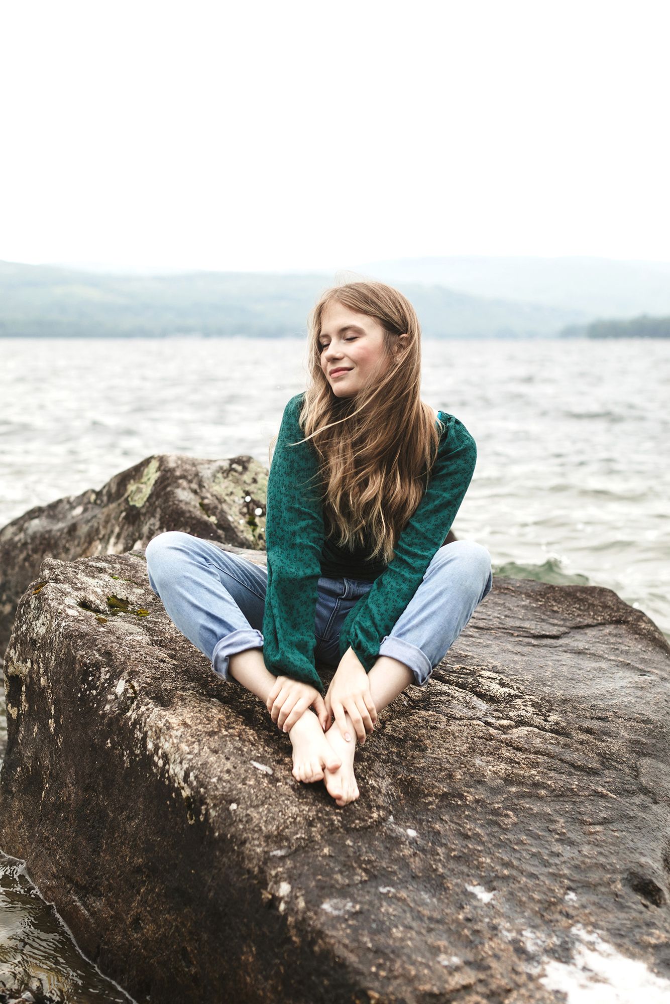 Rachel Delong Christian musician poses on a rock with eyes closed wearing an emerald blouse and light blue jeans at Newfound Lake in NH.