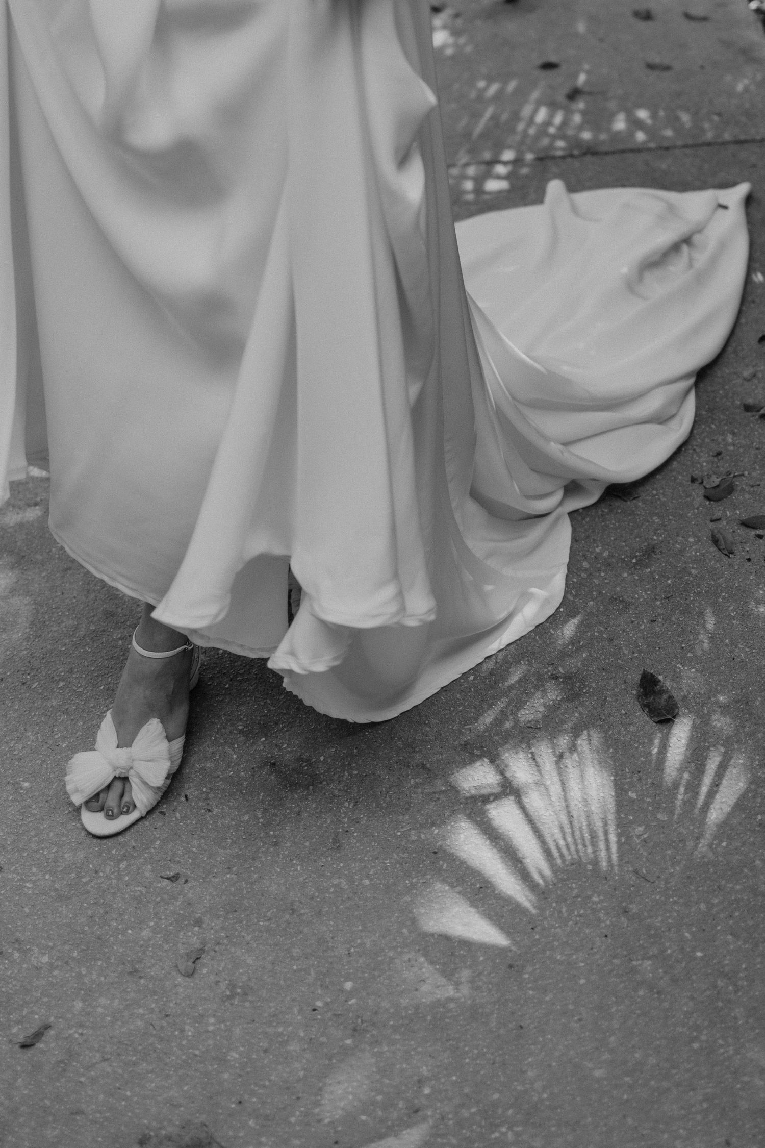 Black and white photo of a bride in a flowing gown wearing bow-detail heels, with palm shadow on ground.
