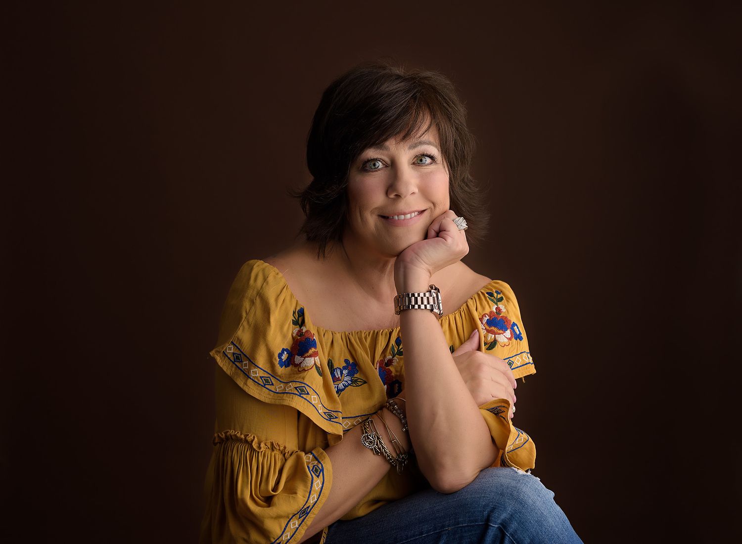 Airdrie photographer with brown hair wearing jeans and yellow shirt looking at camera with chin on hand in Airdrie, Alberta