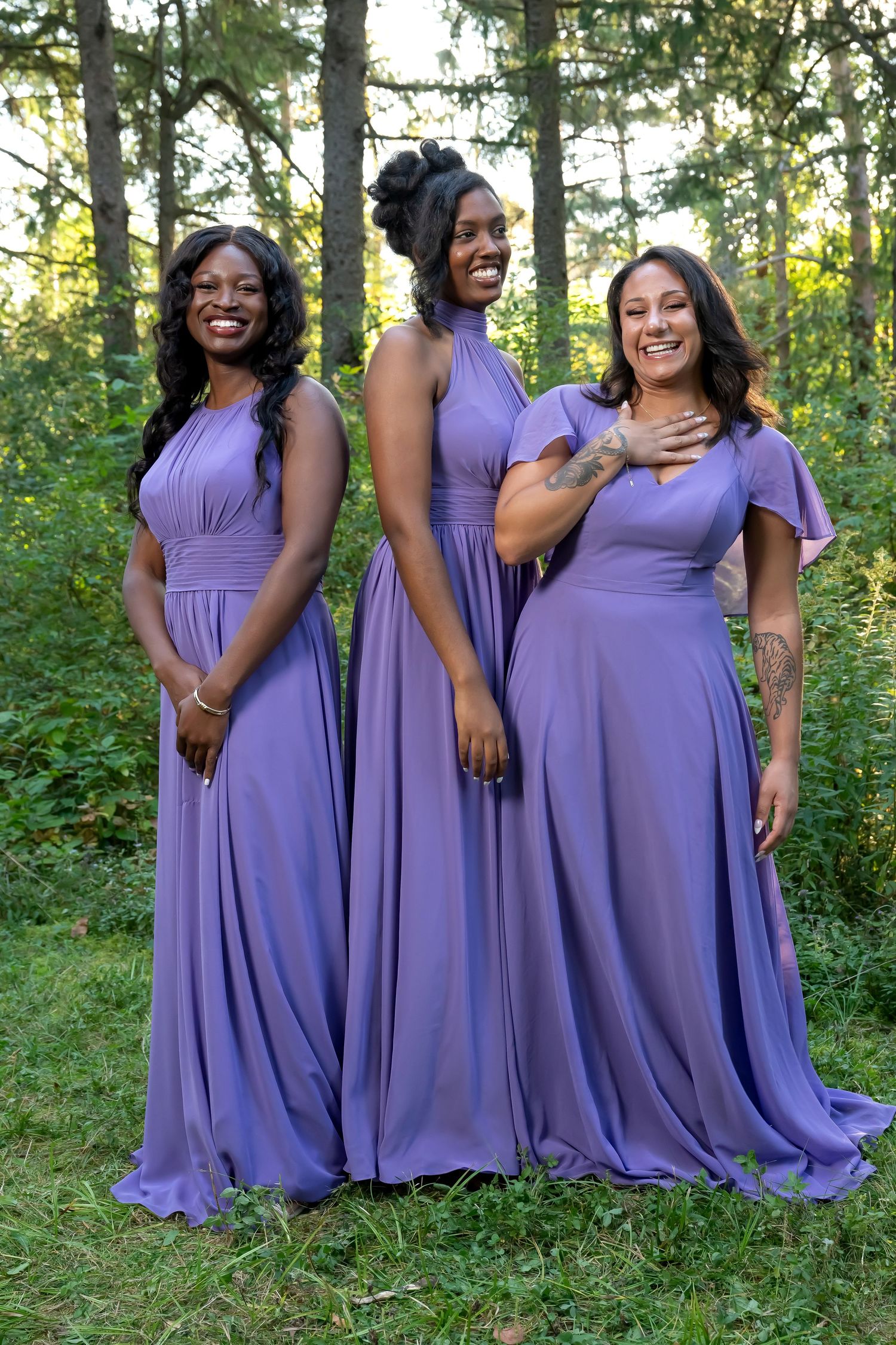 Three bridesmaids wearing matching floor-length lavender dresses pose together in an outdoor woodland setting.