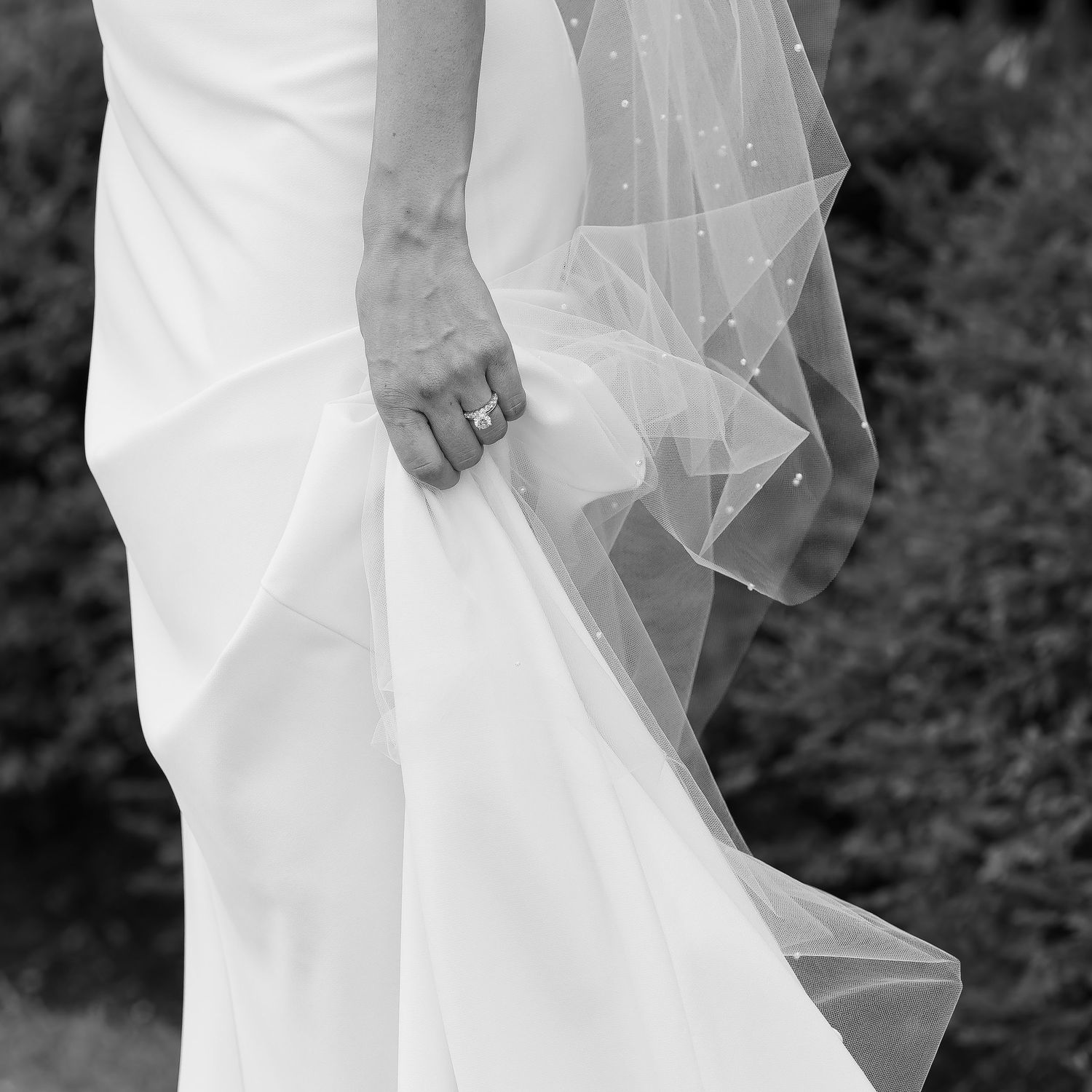 Elegant black and white side view of bride walking, showcasing understated wedding dress, engagement and wedding rings as she holds her gown