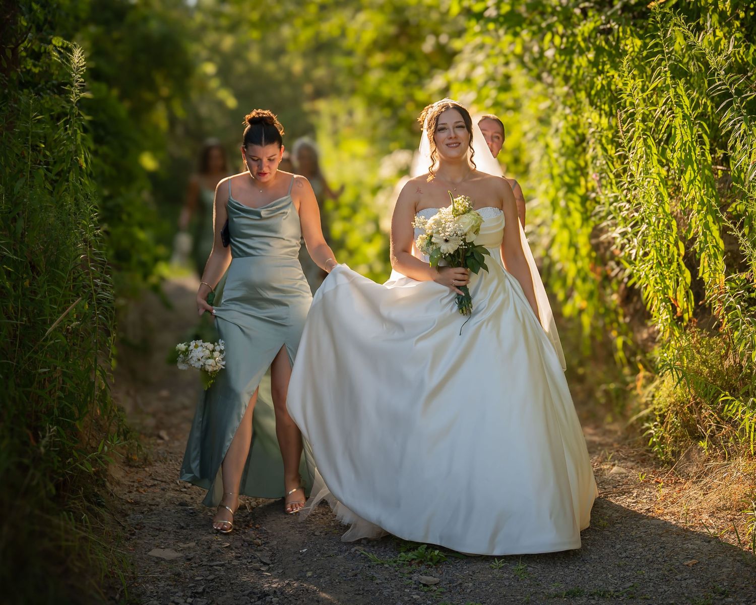 Bride in white gown walks with bridesmaid along a sunlit garden path holding a bouquet.