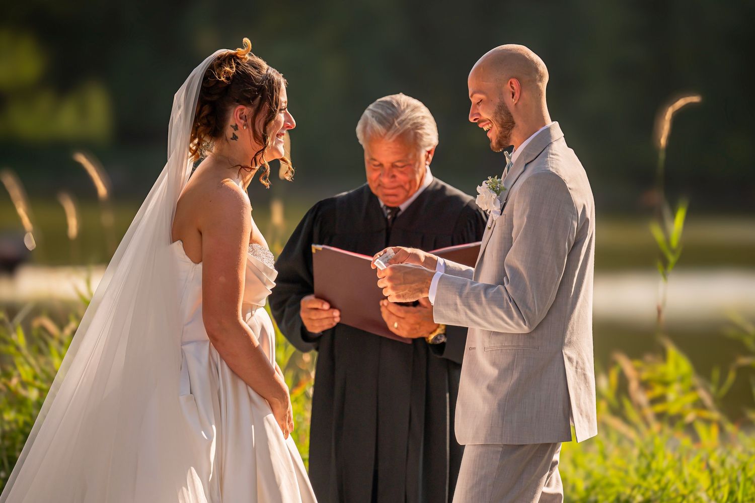 Outdoor wedding ceremony with bride in white gown and groom in gray suit as officiant reads vows by a lake.