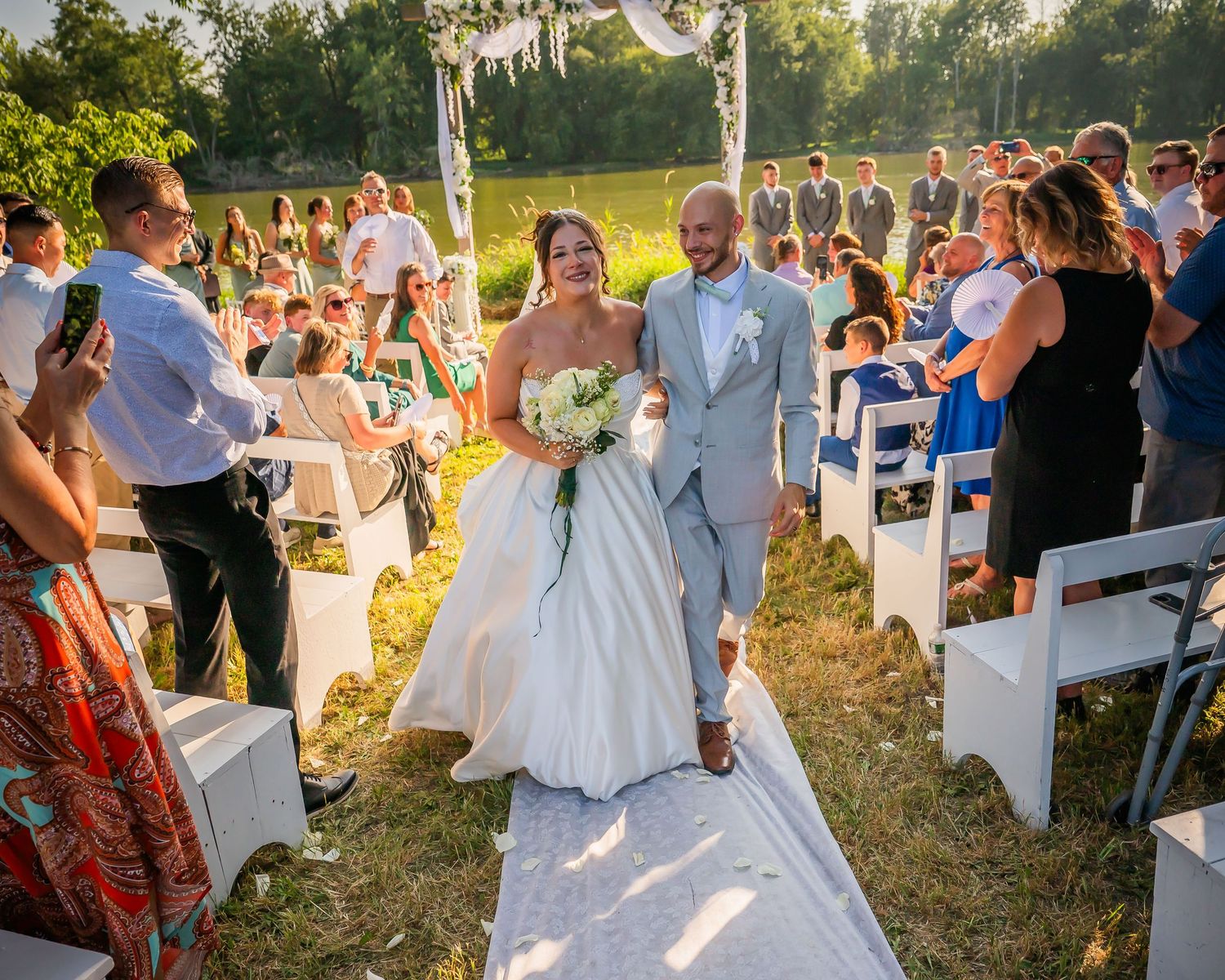 Bride and groom walk down the aisle at an outdoor wedding ceremony surrounded by smiling guests on a sunny day.