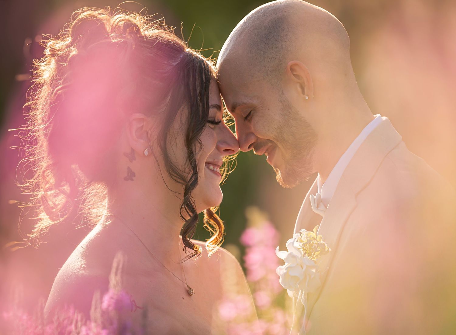 Romantic close-up of smiling wedding couple touching foreheads surrounded by pink flowers in golden light.