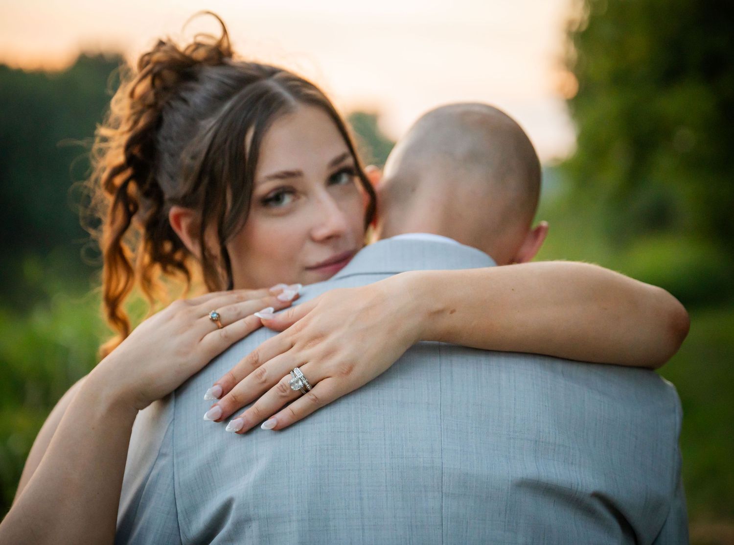 Bride with curly hair hugging groom outdoors at sunset, showcasing her diamond engagement ring.