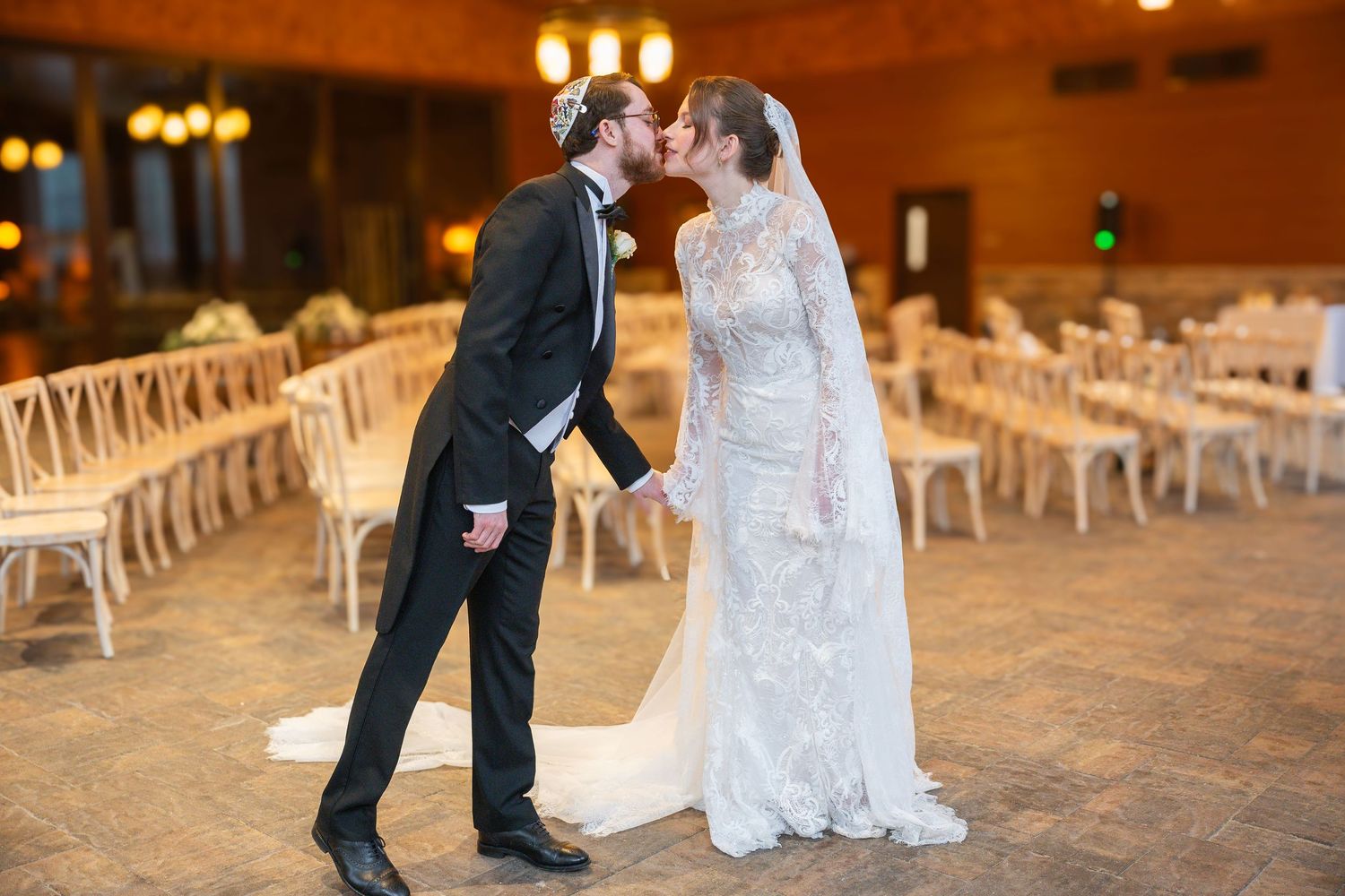 Bride and groom share a romantic kiss in a decorated wedding venue with white chairs and warm ambient lighting.