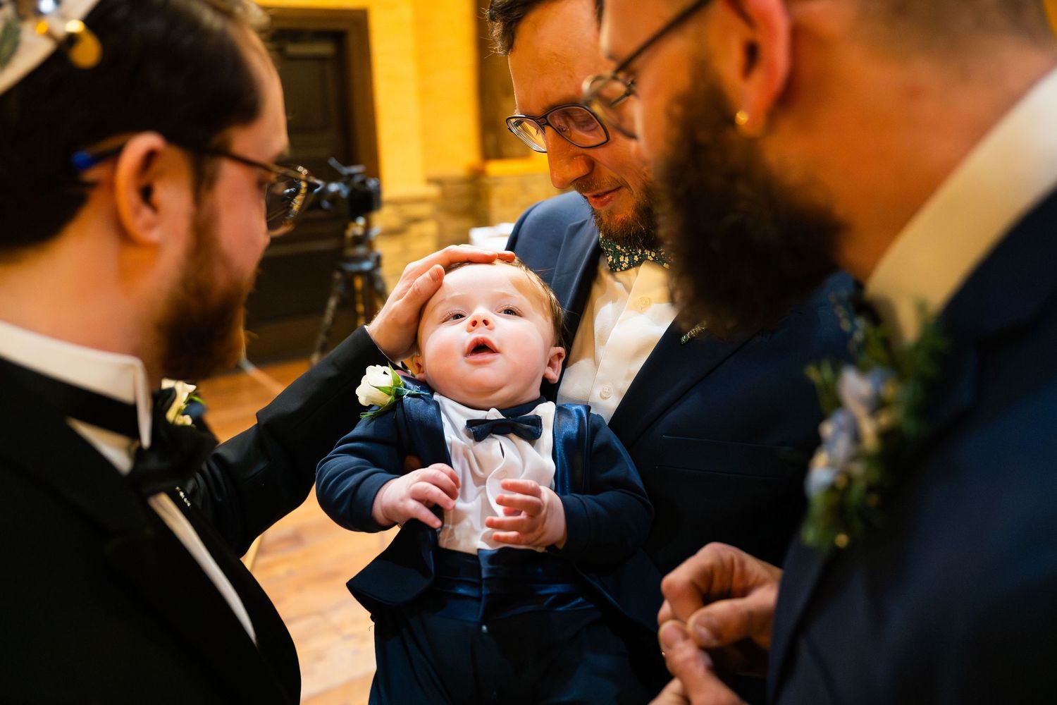 Adorable baby in tiny tuxedo with bow tie held by bearded groomsmen at wedding reception.