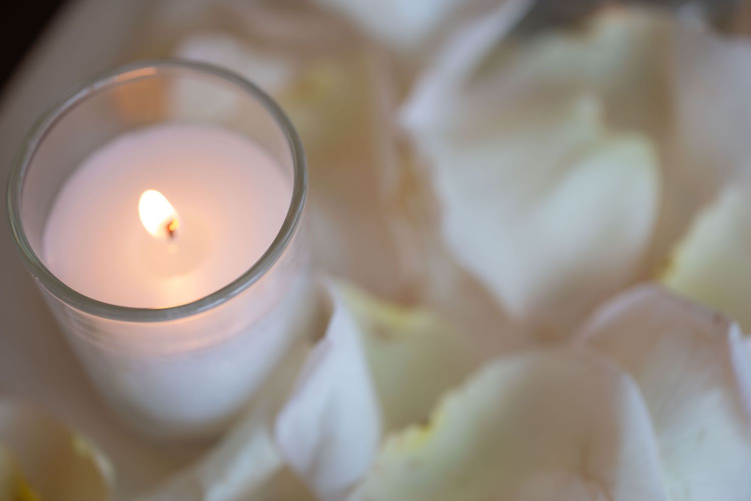 A lit white candle in a glass holder surrounded by soft white rose petals at a wedding.