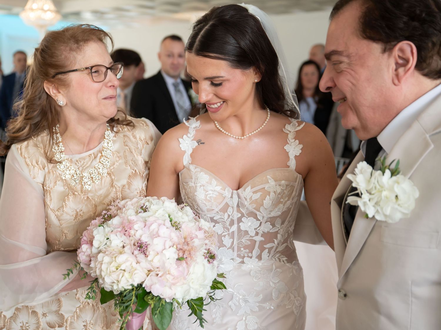 Bride in lace wedding gown holding pink bouquet smiles with parents during wedding ceremony.