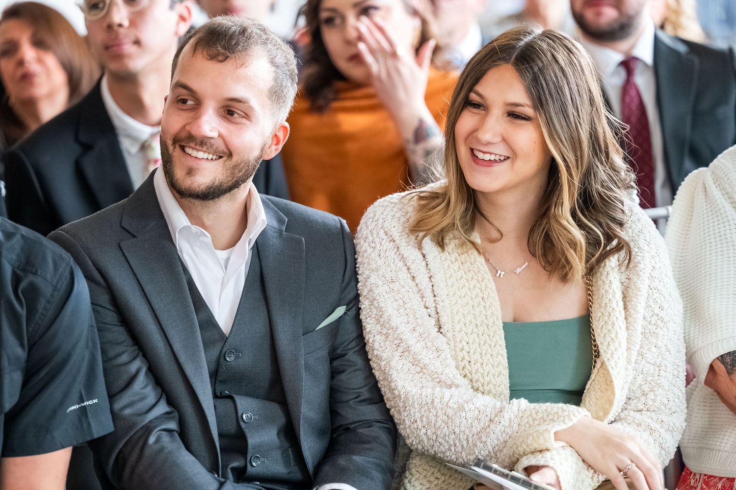 Smiling couple dressed in formal attire sit together as guests at a wedding ceremony.