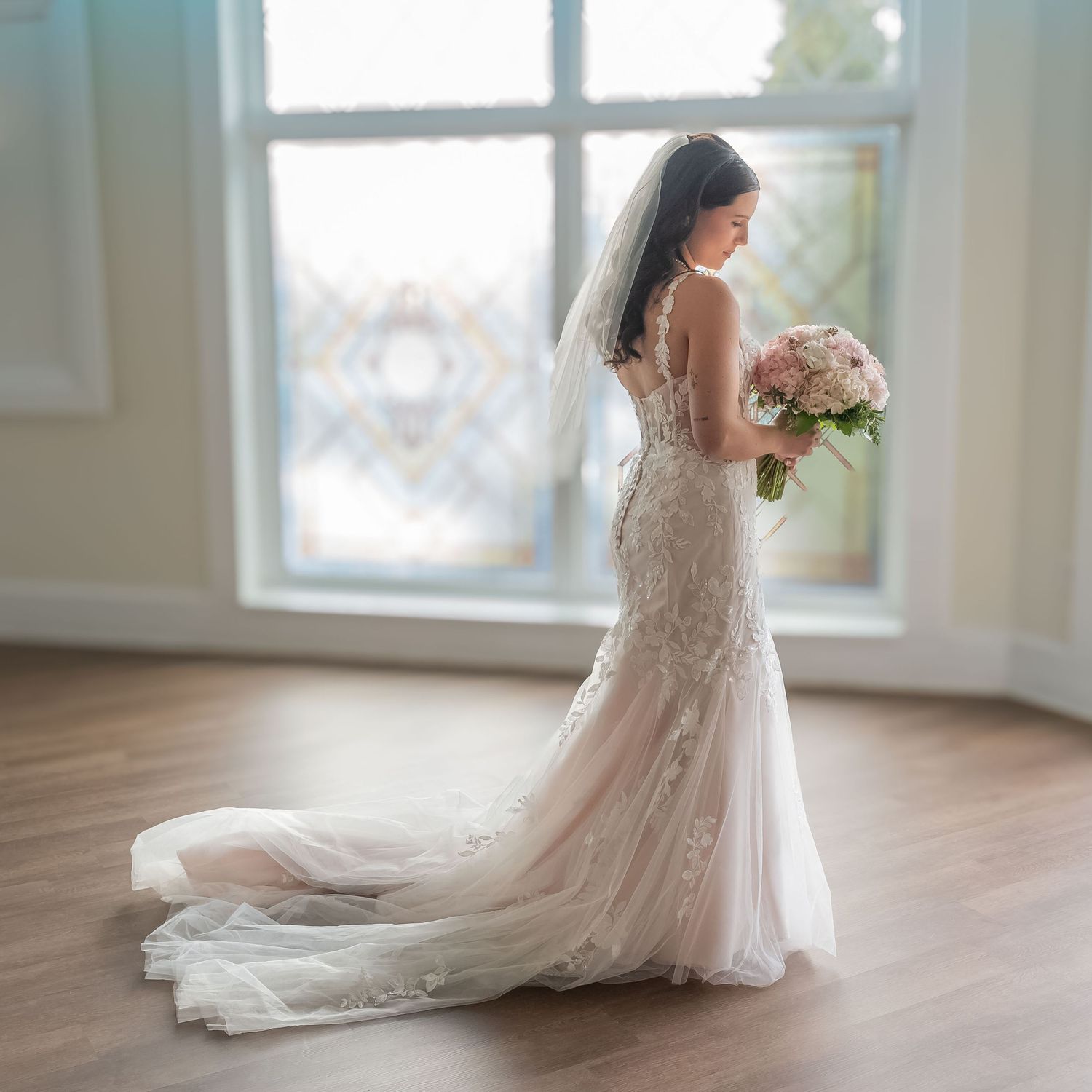 Elegant bride in a lace mermaid wedding dress holding a bouquet standing by a stained glass window.