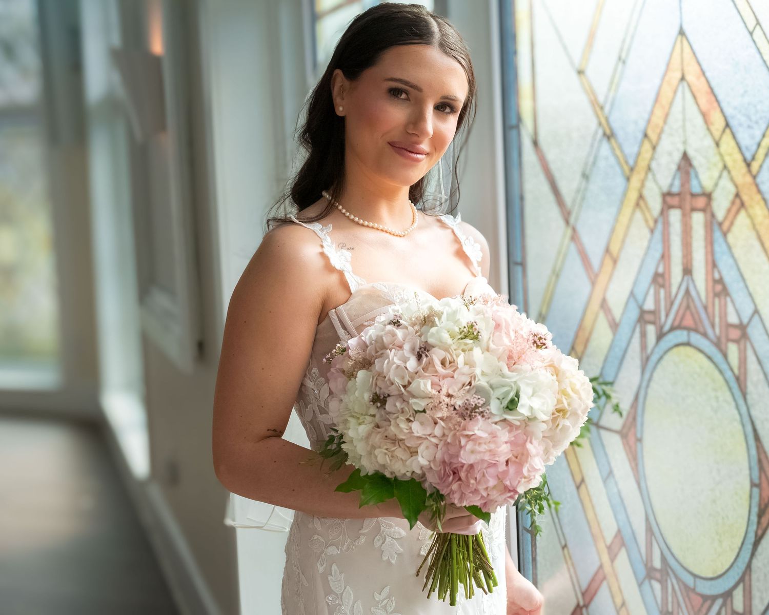 Bride in lace wedding gown holding pink bouquet near stained glass window.