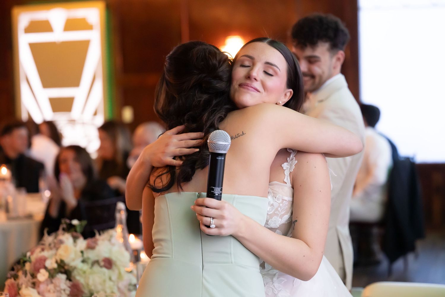 Bridesmaid in sage green dress hugs bride in lace gown during heartfelt wedding reception toast.