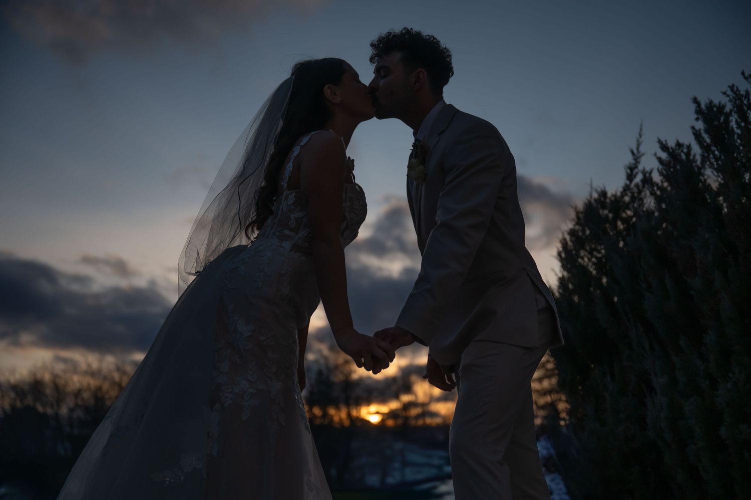 Bride and groom share a romantic kiss at sunset, silhouetted against a dramatic twilight sky at their wedding.
