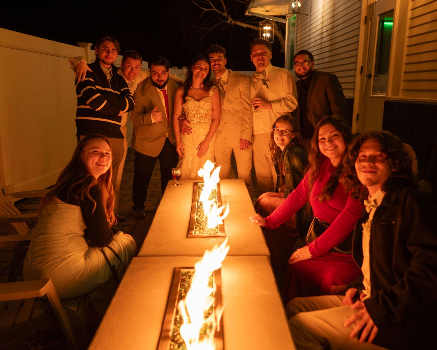 Group of wedding guests gathered around outdoor fire table at night celebrating reception.