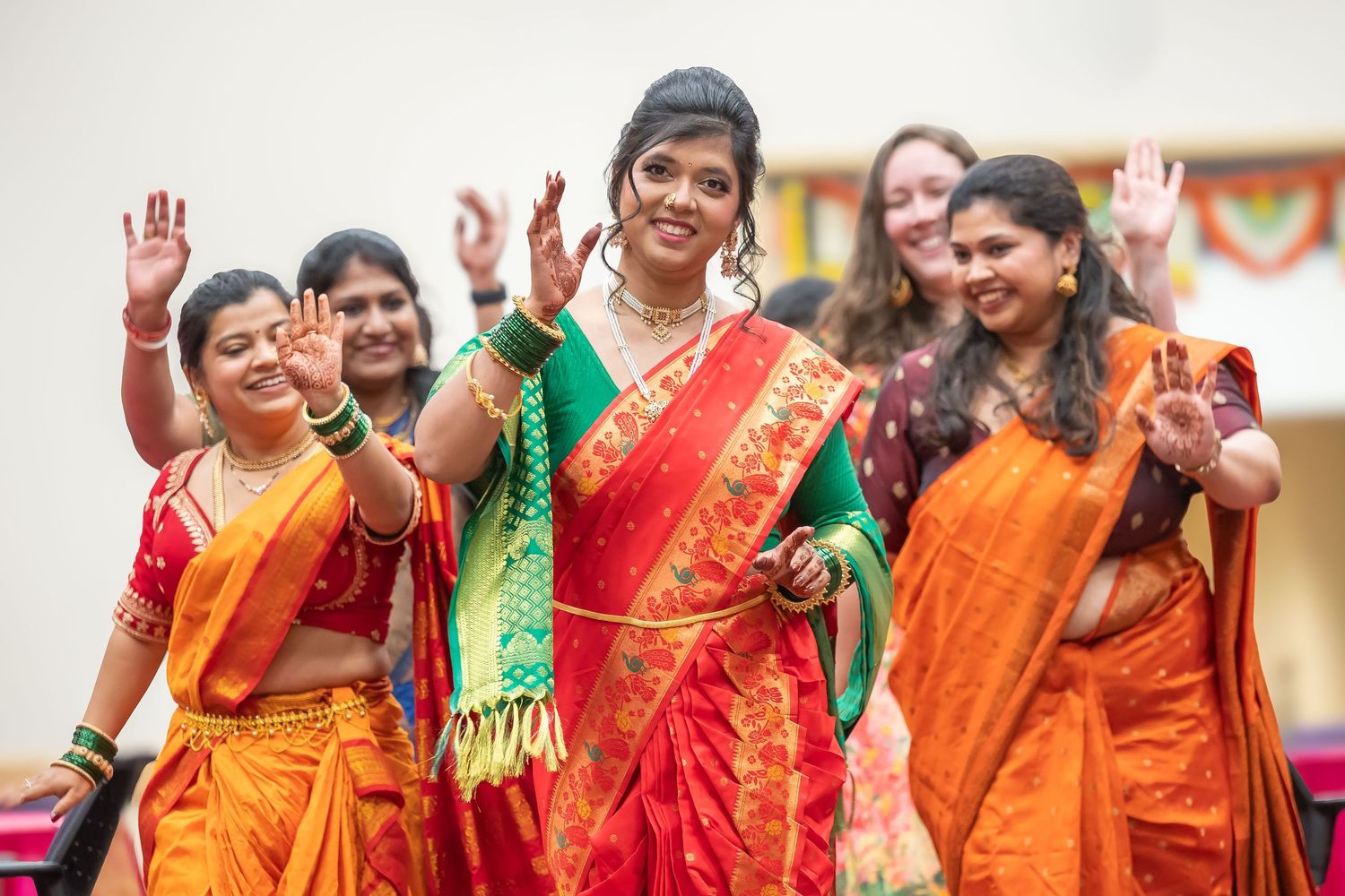 Joyful Indian bride in green and red saree waving with bridesmaids in orange sarees at traditional ceremony.