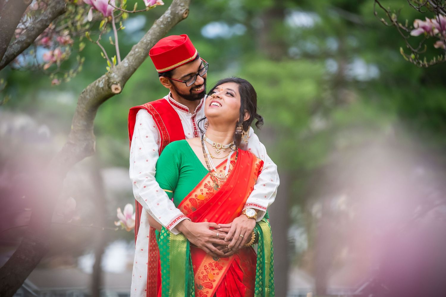 Indian couple in traditional red and green wedding attire embracing under blooming spring trees.