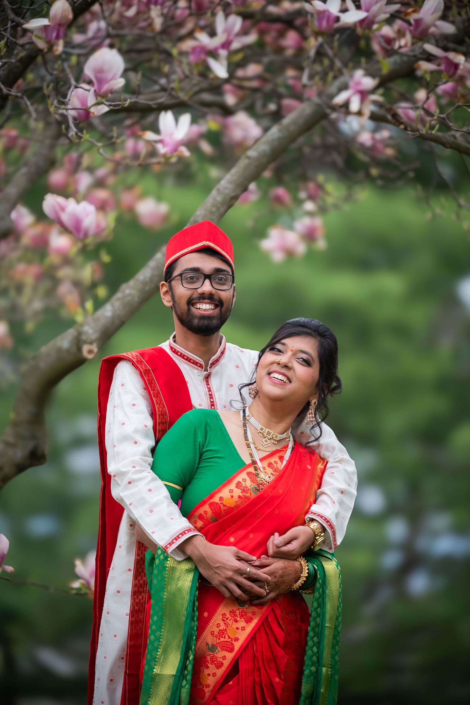 Indian couple in traditional red and green wedding attire posing under blooming magnolia tree.