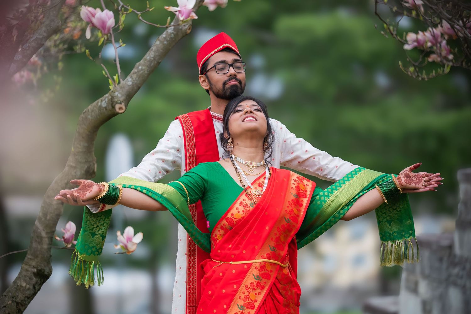 Couple in traditional Maharashtrian wedding attire pose joyfully under blooming magnolia tree.
