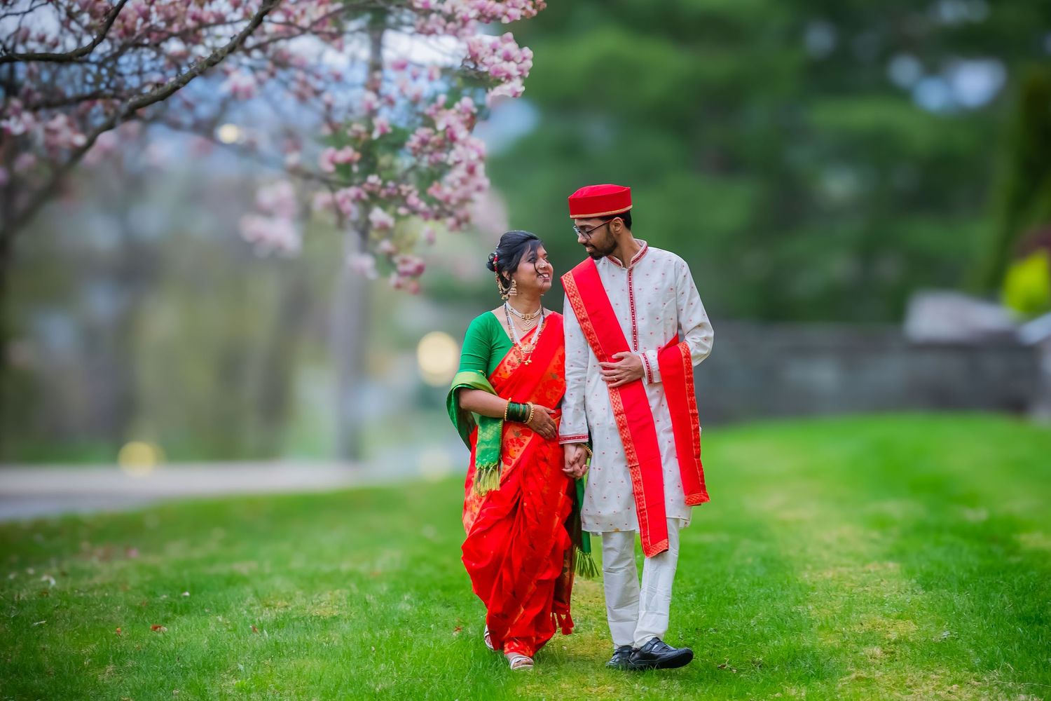 Indian bride in red saree and groom in white kurta walking under pink cherry blossom trees.