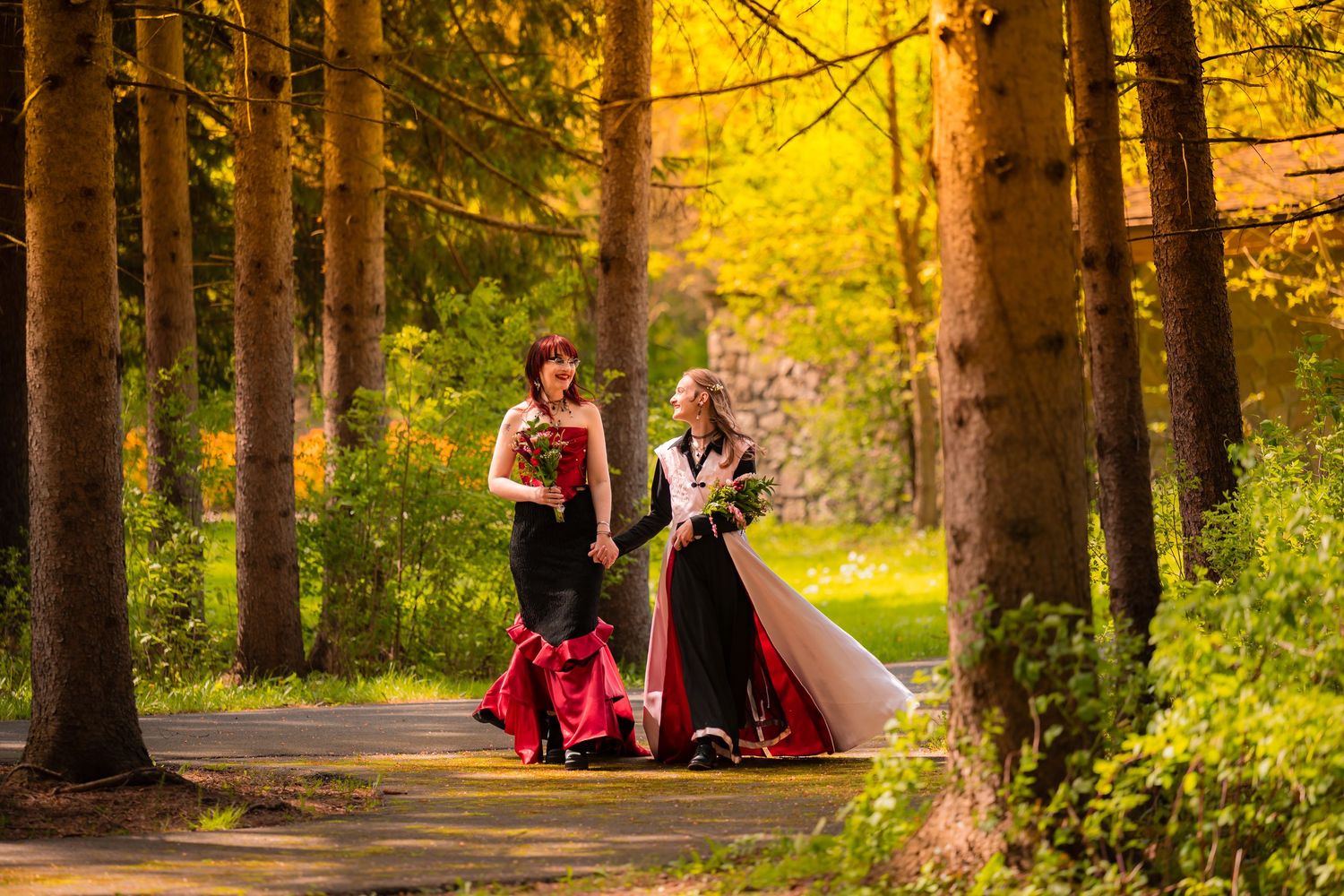 Two women in elegant black and red gowns walk together along a tree-lined path in a lush forest setting.