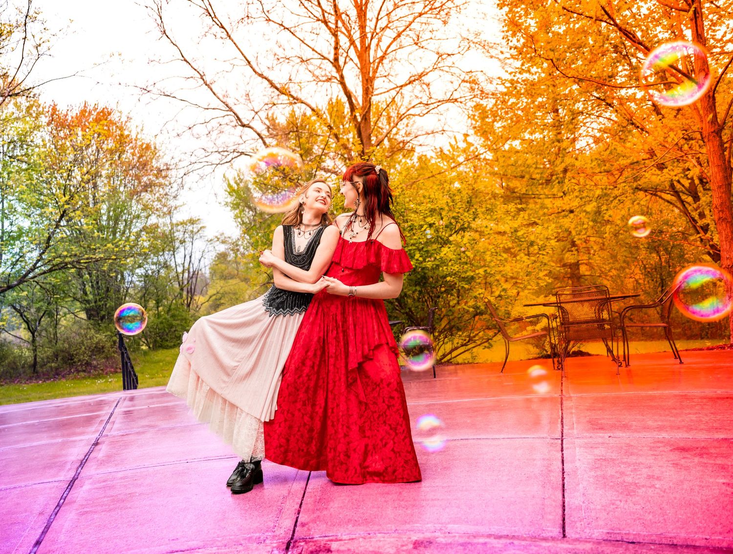 Two women in elegant dresses pose outdoors among autumn trees with colorful soap bubbles floating around them.