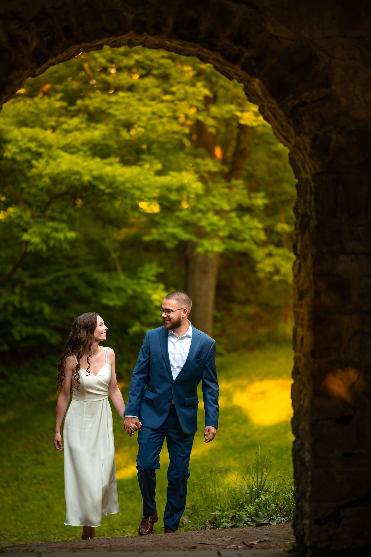 Couple in wedding attire holding hands walking through a stone arch surrounded by lush green trees.