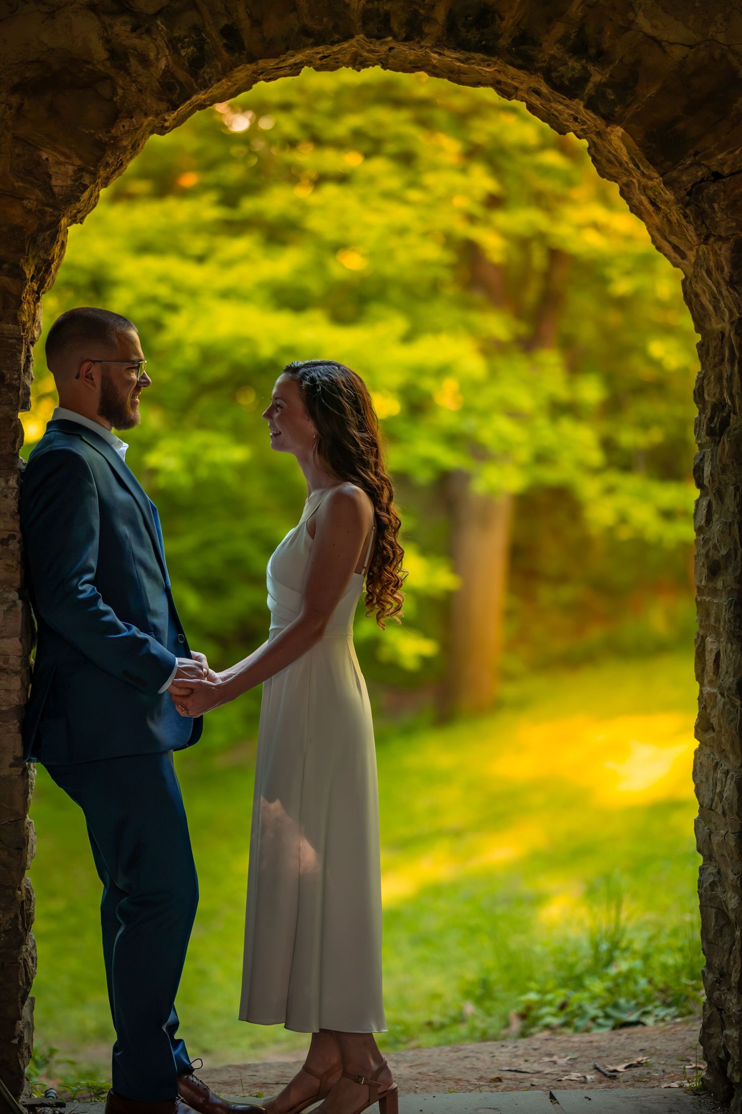 Bride and groom hold hands under stone arch framed by lush green forest at golden hour.