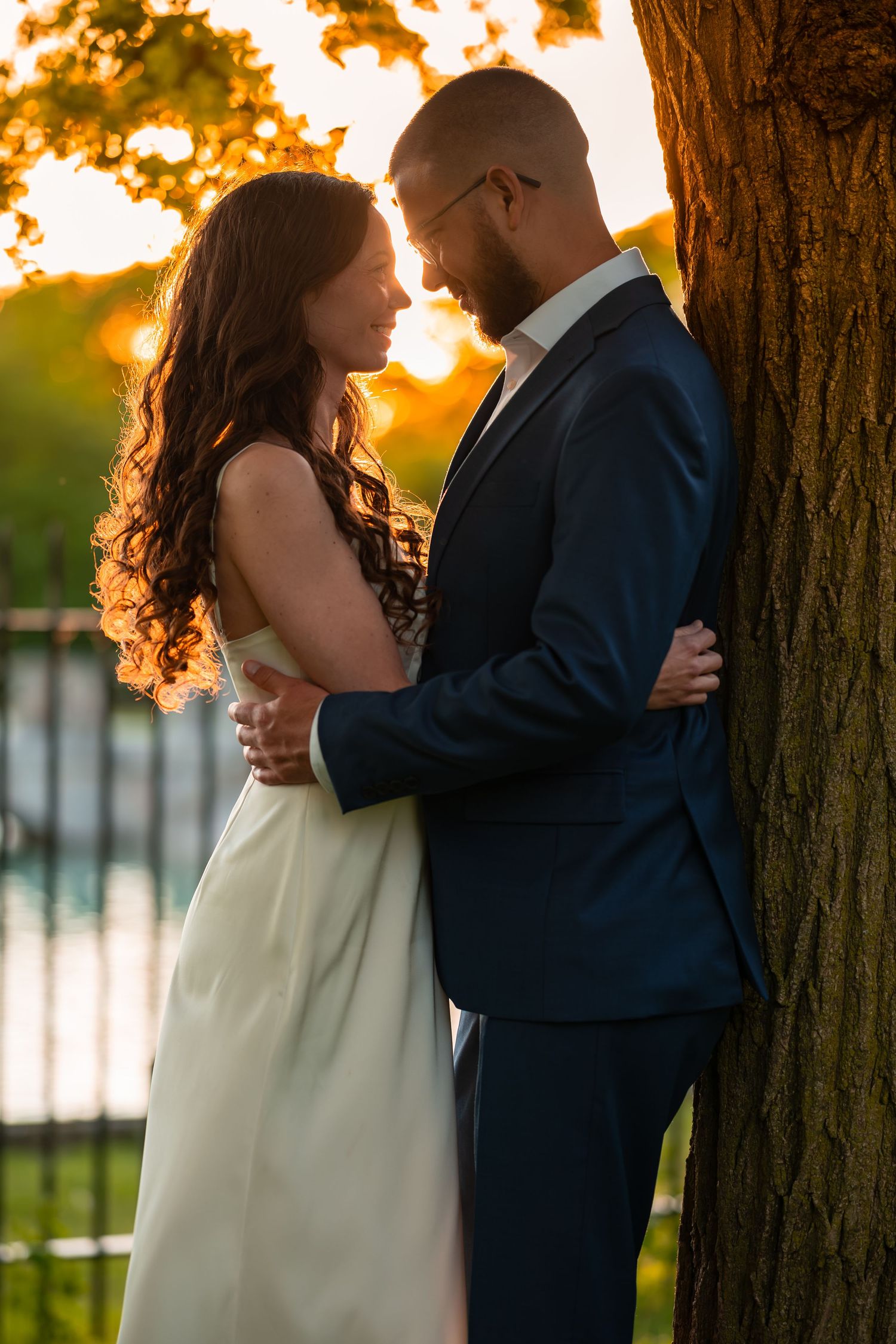 Bride and groom embracing by tree trunk in romantic golden sunset light near water.