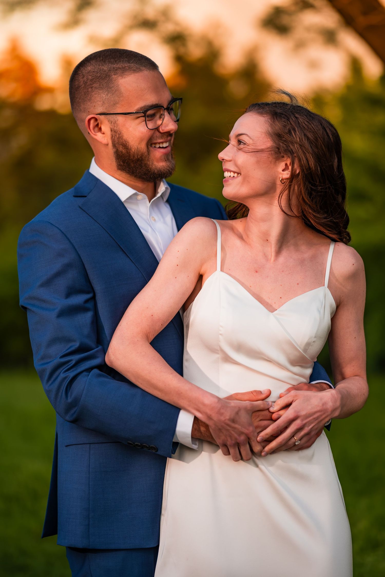 A smiling groom in a blue suit embraces his bride in a white slip dress during a golden hour outdoor shoot.
